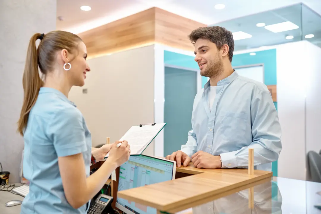A receptionist with a clipboard welcoming a patient at a clinic’s front desk.