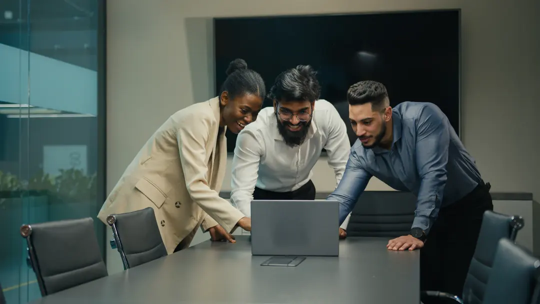 Three colleagues gathered round a laptop in a glass-walled meeting room.