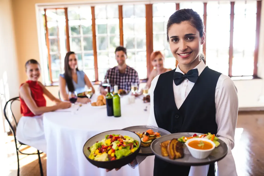 A server carrying plates through a full restaurant, a table of guests behind her.