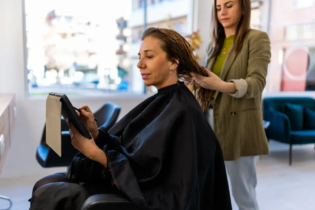 A stylist working on a client’s hair in a bright, modern salon while the client checks a tablet.